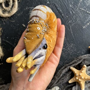 Może przedstawiać: Pluszowy nautilus ze złotą muszlą i mackami. Muszla jest ozdobiona srebrnymi i złotymi koralikami. Nautilus jest trzymany w dłoni.