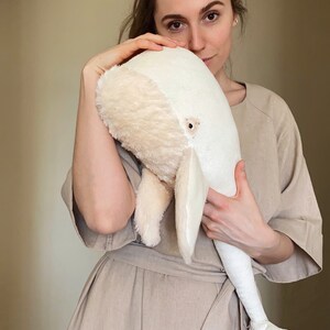 Puede incluir: Una mujer sostiene un gran juguete de peluche de ballena blanca. La ballena es suave y esponjosa, con una textura peluda. La mujer lleva un vestido marr&oacute;n claro.