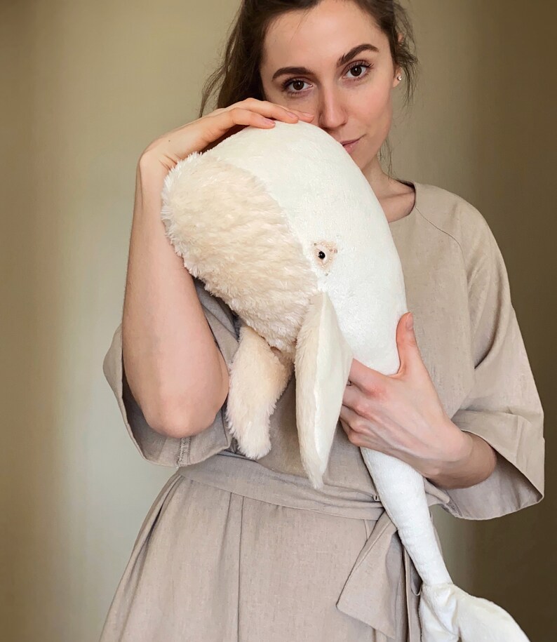 Puede incluir: Una mujer sostiene un gran juguete de peluche de ballena blanca. La ballena es suave y esponjosa, con una textura peluda. La mujer lleva un vestido marr&oacute;n claro.