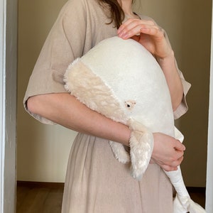 Puede incluir: Un juguete de peluche de ballena blanca con una textura suave y esponjosa. La ballena est&aacute; siendo sostenida por una persona que lleva un vestido beige.