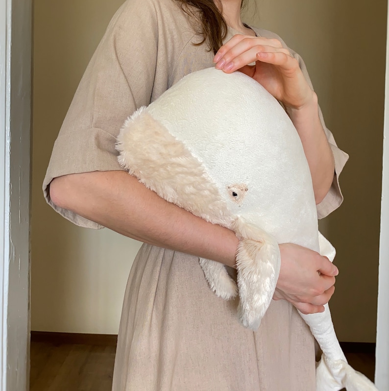 Puede incluir: Un juguete de peluche de ballena blanca con una textura suave y esponjosa. La ballena est&aacute; siendo sostenida por una persona que lleva un vestido beige.