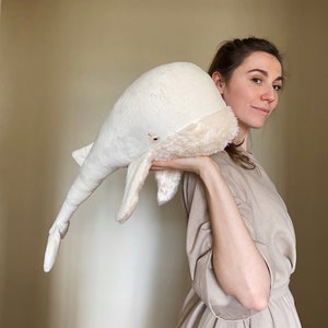 Puede incluir: Una mujer con un vestido beige sostiene un gran juguete de ballena de peluche blanco frente a ella.
