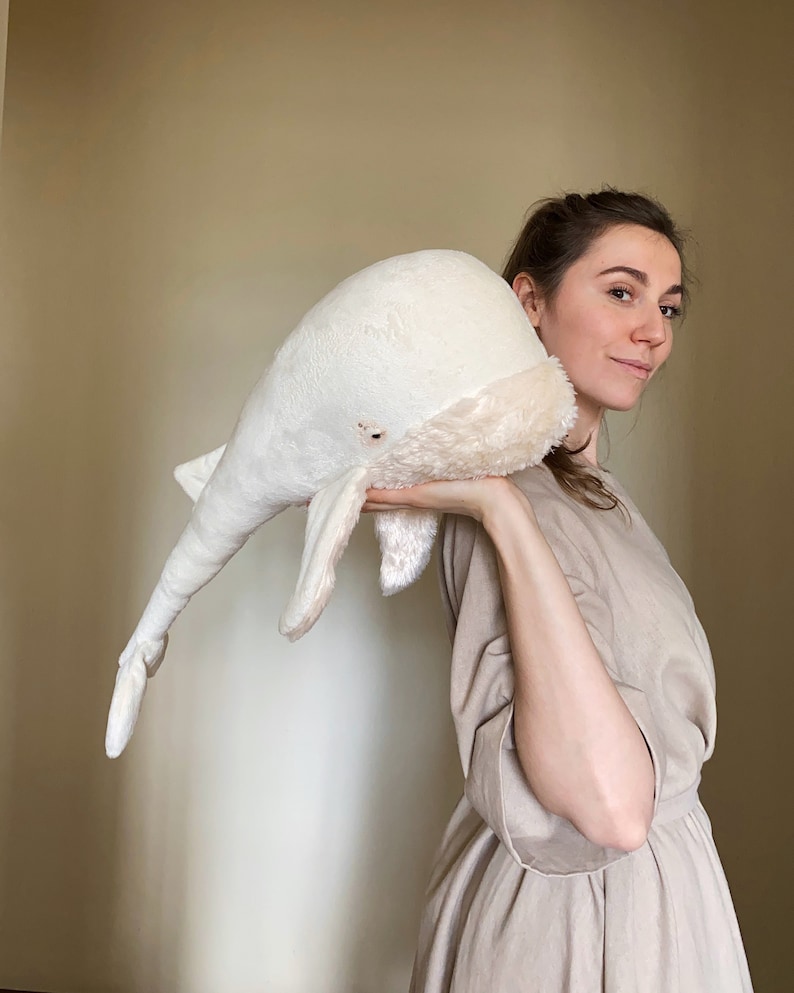 Puede incluir: Una mujer con un vestido beige sostiene un gran juguete de ballena de peluche blanco frente a ella.