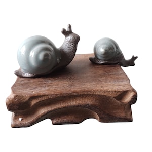 May include: Two ceramic snail figurines on a wooden base. The larger snail is light blue and the smaller snail is dark grey.