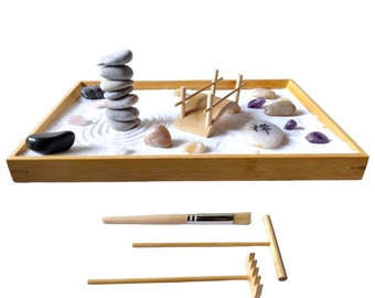 May include: A wooden tray with white sand, a small wooden bridge, grey stones, and purple and pink crystals. The tray also includes a rake and a brush for creating patterns in the sand. The tray has the word "Zen" written on a stone.