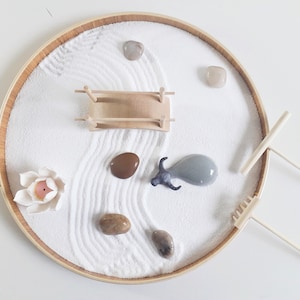 May include: A white sand zen garden set in a round wooden tray. The set includes a small wooden bridge, a grey ceramic bull figurine, a white ceramic lotus flower, and two wooden rakes. There are also several small stones in the sand.