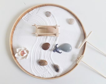 May include: A white sand zen garden set in a round wooden tray. The set includes a small wooden bridge, a grey ceramic bull figurine, a white ceramic lotus flower, and two wooden rakes. There are also several small stones in the sand.