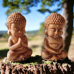 May include: Two small wooden statues of Buddha sitting in meditation pose. The statues are carved from light brown wood and have closed eyes. They are sitting on a mossy tree stump.