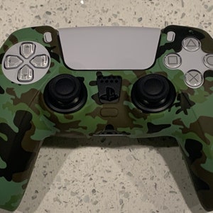 Puede incluir: Un mando de juego PlayStation 5 DualSense de color verde y negro camuflaje con botones blancos y un panel táctil blanco.