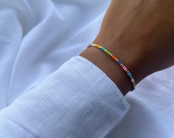 May include: A colorful beaded bracelet with gold accents. The bracelet is made with small, round beads in various colors, including pink, blue, green, yellow, and orange. The beads are strung on a thin gold chain.