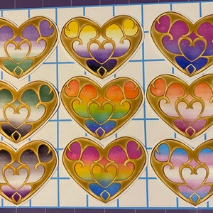 May include: Nine heart-shaped stickers with gold filigree borders and colorful, gradient-filled interiors. The stickers feature various pride flag color schemes, including rainbow, purple, and blue, on a grid background. Each heart has a double-heart design.