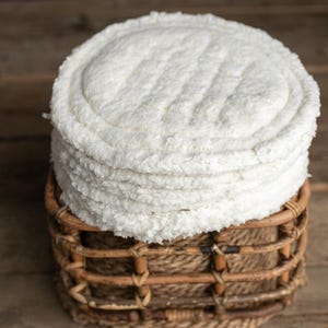 May include: A white, fluffy, oval-shaped cushion sits in a woven wicker basket. The cushion is made of a soft, plush material and appears to be made of several layers.