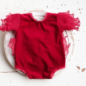 May include: A red velvet baby bodysuit with ruffled sleeves. The sleeves are made of a sheer fabric.