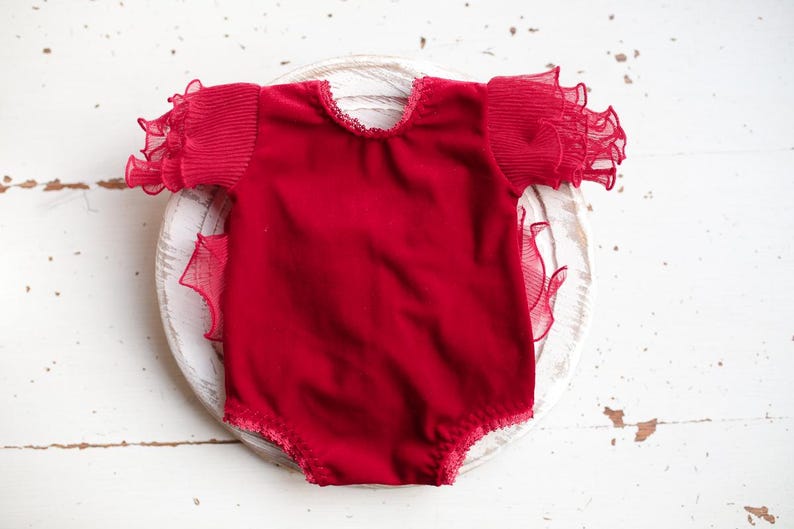May include: A red velvet baby bodysuit with ruffled sleeves. The sleeves are made of a sheer fabric.