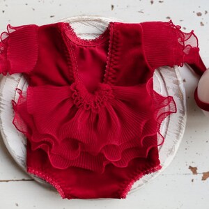 May include: Red baby outfit with a ruffled skirt and short sleeves. The outfit is made of a soft, velvet-like fabric and has a delicate lace trim.