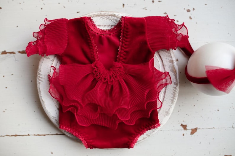 May include: Red baby outfit with a ruffled skirt and short sleeves. The outfit is made of a soft, velvet-like fabric and has a delicate lace trim.