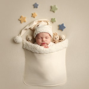 Pode incluir: Um recém-nascido usando um chapéu de tricô branco dorme em um envoltório branco com uma borda branca felpuda. O bebê está cercado por pequenas estrelas de feltro em tons de azul, verde e amarelo. Dois pequenos animais de pelúcia são visíveis perto da cabeça do bebê.