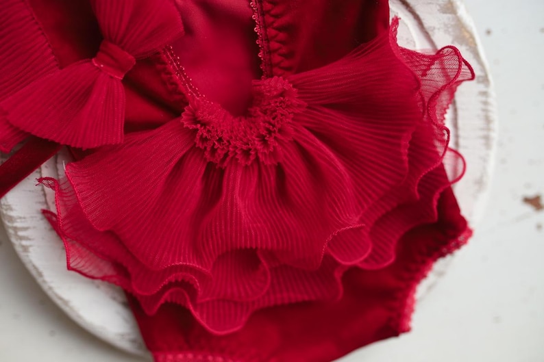 May include: Red ruffled baby bodysuit with a large bow on the front. The bodysuit is made of a soft, delicate fabric and is perfect for a special occasion.