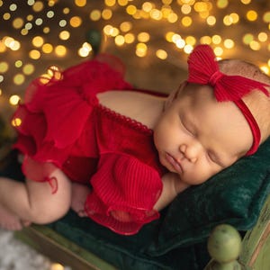 May include: A newborn baby girl sleeping on a green velvet pillow in a red velvet dress with a red bow headband. The baby is surrounded by warm, golden lights.