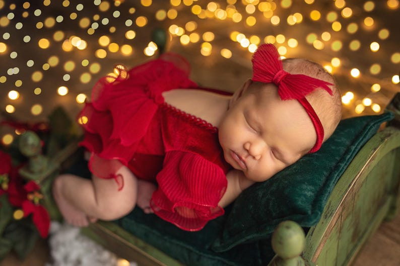 May include: A newborn baby girl sleeping on a green velvet pillow in a red velvet dress with a red bow headband. The baby is surrounded by warm, golden lights.