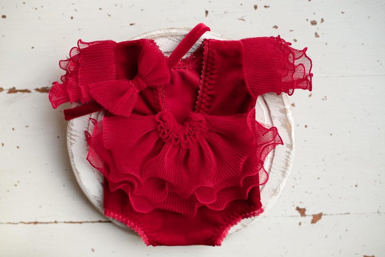 May include: Red ruffled baby outfit with a matching headband. The outfit has a layered ruffle design and a bow on the front.