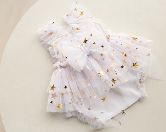 Op de afbeelding: Wit tule babyjurk met gouden sterrendruk. De jurk heeft een gerimpelde halslijn en een gelaagde rok.