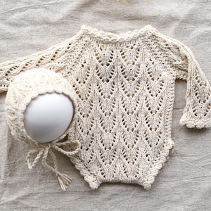 Pode incluir: Um conjunto de bebé em crochê de cor creme com mangas compridas e um gorro a condizer. O conjunto tem um padrão delicado de ponto aberto.
