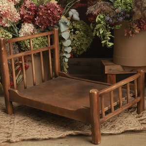 Puede incluir: Una pequeña cama de madera rústica con cabecero y piecero, ideal para fotografía de recién nacidos. La cama marrón se encuentra sobre una alfombra de yute tejida. Flores secas y vegetación en el fondo.