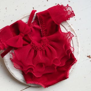 May include: Red baby outfit with a large bow and ruffled trim. The outfit is made of a soft, velvet-like fabric.
