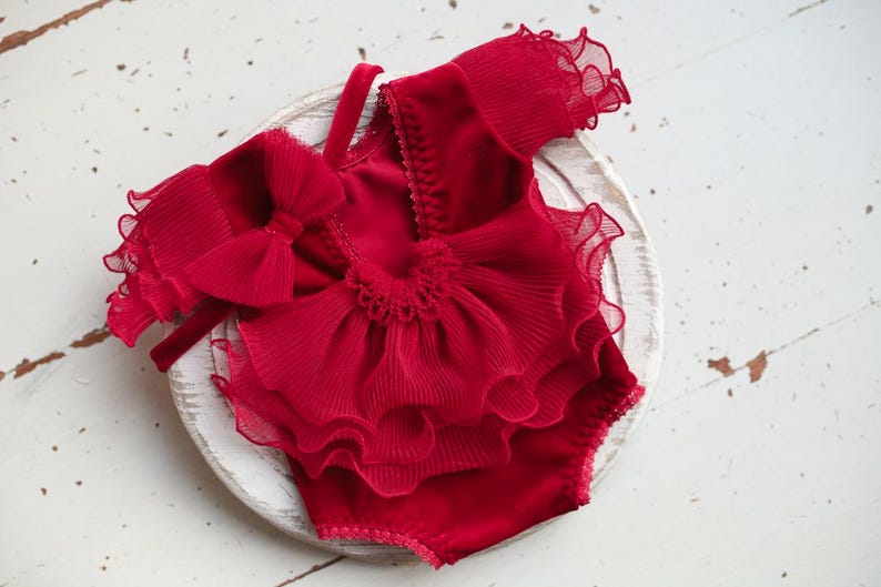May include: Red baby outfit with a large bow and ruffled trim. The outfit is made of a soft, velvet-like fabric.