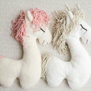 May include: Two plush unicorn toys. One has a pink mane and tail, the other has a beige mane. Both are white with black stitched eyes and a stitched mouth. The toys are lying on a textured, off-white surface.