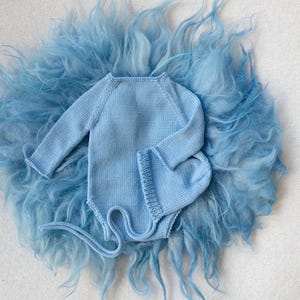 May include: A light blue knitted baby bodysuit and matching hat are displayed on a fluffy, light blue faux fur background. The bodysuit has long sleeves and a rounded neckline. The hat is a beanie style. The set is designed for newborns.
