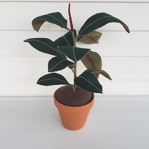 May include: A handcrafted felt plant in a terracotta pot. The dark green leaves have white stitching and red accents. The plant sits on a brown base inside the pot. The pot is a light orange color.