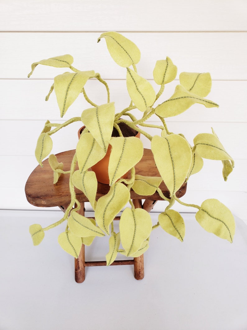 May include: A felt plant with green leaves in a terracotta pot, sitting on a wooden stool. The plant is hanging over the sides of the stool.