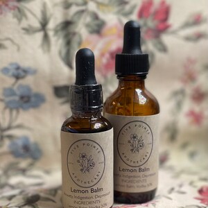 May include: Two amber glass bottles with black dropper tops and brown paper labels. The labels have a floral design and the text "Five Points Apothecary" and "Lemon Balm". The smaller bottle lists the ingredients as "Lemon Balm, Vodka 50%". The larger bottle lists the ingredients as "Lemon Balm, Vodka 10%".