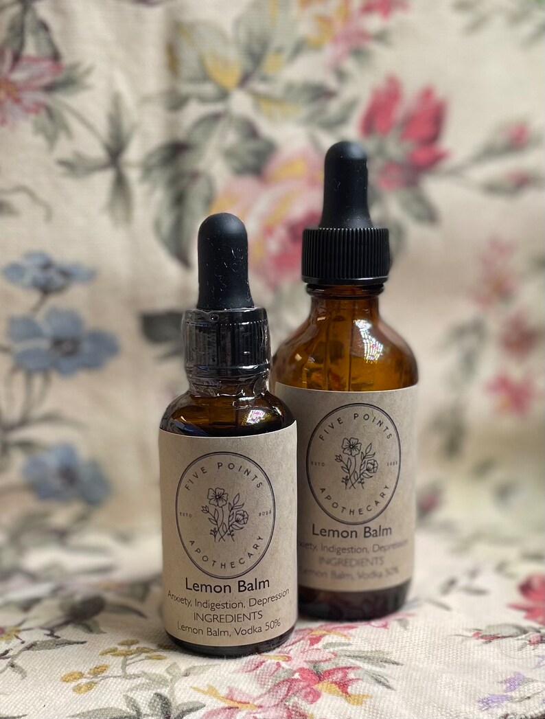May include: Two amber glass bottles with black dropper tops and brown paper labels. The labels have a floral design and the text "Five Points Apothecary" and "Lemon Balm". The smaller bottle lists the ingredients as "Lemon Balm, Vodka 50%". The larger bottle lists the ingredients as "Lemon Balm, Vodka 10%".