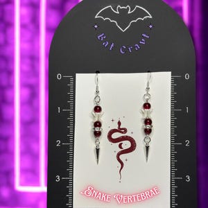 May include: A pair of dangle earrings with a gothic theme. The earrings feature white bone-shaped beads, red beads, and silver-colored metal accents. The earrings hang from a black display card with the text "Bat Cravt" and "Snake Vertebrae".