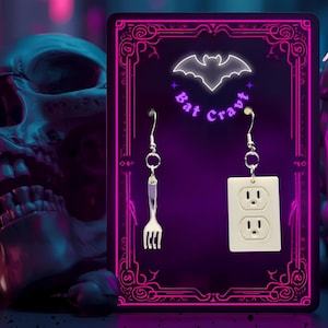 May include: A pair of novelty earrings displayed on a purple background. One earring is a silver fork, the other is a white electrical outlet. The display card has a neon pink border and the text "Bat Cravt" with a bat symbol.