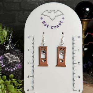May include: A pair of brown guillotine earrings with silver-colored metal hooks are displayed on a white stand. The stand has a bat logo and the text "Bat Cravt". The earrings are a unique accessory.