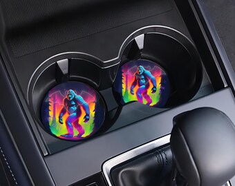 May include: Two round car coasters with a colorful, retro-style illustration of a large, blue gorilla standing in a forest. The coasters are in a car's cup holder.