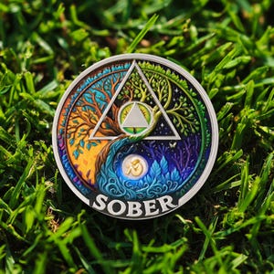 Könnte beinhalten: Eine runde, silberfarbene Münze mit einem farbenfrohen Design, das einen Baum, ein Dreieck und ein Yin-Yang-Symbol zeigt. Das Wort "SOBER" ist unten eingraviert. Die Münze liegt auf grünem Gras.