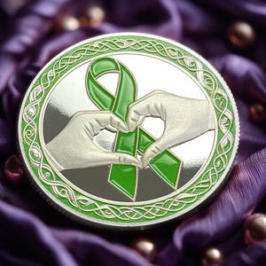 May include: A silver coin with a green ribbon tied in a bow and two hands forming a heart around it. The coin has a Celtic knot design around the edge.