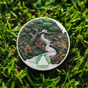 Può includere: Una moneta rotonda color argento con un rilievo dettagliato di un albero, un fiume e fiori. La moneta ha un triangolo verde e bianco con le parole "Unity, Service, Recovery". La moneta è posta su uno sfondo di erba verde.