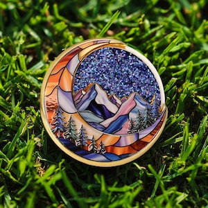 May include: A round, gold-toned pin with a mountain and forest scene. The design features a crescent moon shape with stained glass-style panels in orange, purple, and blue. The center has a glittery blue starry sky.