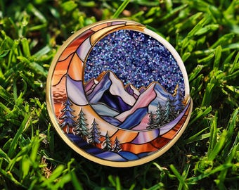 May include: A round, gold-toned pin with a mountain and forest scene. The design features a crescent moon shape with stained glass-style panels in orange, purple, and blue. The center has a glittery blue starry sky.