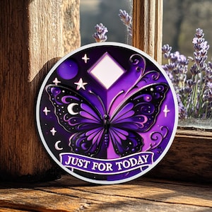 Pode incluir: Um autocolante redondo com um desenho de borboleta em tons de roxo e preto. A borboleta tem padrões intrincados nas asas e está centrada no autocolante. O texto "JUST FOR TODAY" é exibido numa faixa na parte inferior. Estrelas, luas e um losango também estão incluídos.