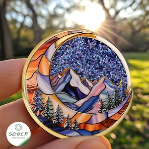 May include: A round, gold-toned pin featuring a mountain landscape under a starry night sky, framed by a crescent moon. The design incorporates shades of orange, purple, blue, and green, with a glittery center. The pin is held in a hand.