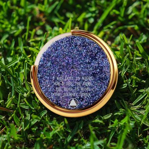 May include: A round, gold-colored phone grip with a glittery, purple center. The grip has the text "WAS LOST IN NIGHT, NOW I SEE THE DAWN. THE ROAD IS MINE, THE JOURNEY SOBER." The phone grip is on a bed of green grass.