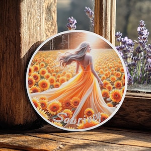 May include: A round, silver-coloured coin featuring a colourful image of a woman in a yellow dress walking through a field of sunflowers. The word "Sobriety" is written in cursive at the bottom of the coin.