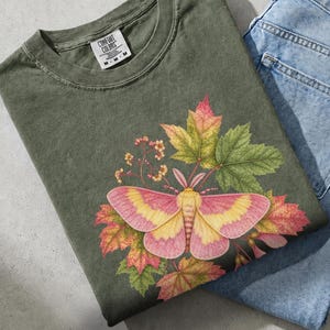 May include: A moss green short-sleeve t-shirt featuring a graphic of a pink and yellow moth surrounded by autumn-coloured maple leaves and small flowers. The shirt has a Comfort Colors brand label at the neck and is folded next to blue denim jeans.
